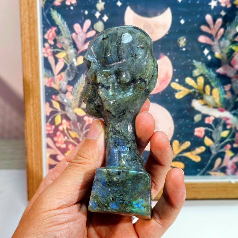 Puede incluir: Una escultura de busto de labradorita tallada. La escultura es una cabeza y cuello sobre una base rectangular. La piedra tiene una mezcla de colores verdes, azules y grises. El busto est&aacute; colocado contra un fondo enmarcado con elementos florales y celestiales.