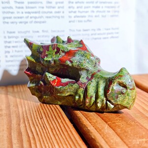 May include: A carved dragon head sculpture in shades of green and red. The sculpture is made of a stone-like material and has detailed features. The dragon head is resting on a wooden surface.