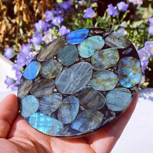 May include: An oval-shaped decorative piece featuring a mosaic of polished labradorite stones in shades of blue, teal, and gray. The stones are set in a dark, possibly black, matrix. The piece is held in a hand, with a blurred floral background.