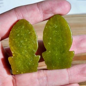 Puede incluir: Dos colgantes de jade verde con una forma &uacute;nica, con una parte superior redondeada y una base acampanada. El jade tiene un aspecto moteado con variaciones de color, que van del verde claro al oscuro.