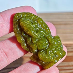 Puede incluir: Un colgante de jade verde tallado en forma de deidad. El colgante tiene detalles intrincados y un acabado pulido. El jade es de un color verde vibrante, y el tallado representa una figura con rasgos ornamentados. El colgante se sostiene en una mano.