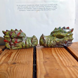 May include: Two carved stone dragon heads. The heads are green with red accents and are placed on a wooden surface. The text in the background reads: "In my opinion, lovers not simply mean your partner of life..."