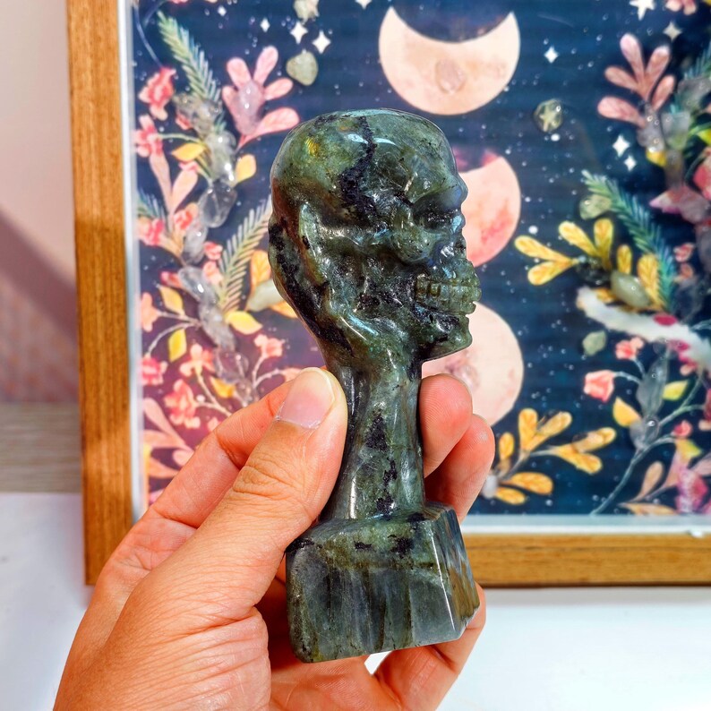 Puede incluir: Una figura de calavera tallada, de color verde, con un rostro esquel&eacute;tico detallado. La escultura se sostiene en una mano, mostrando su intrincado dise&ntilde;o y superficie pulida. La base es rectangular y el fondo presenta una obra de arte enmarcada con elementos celestiales y florales.