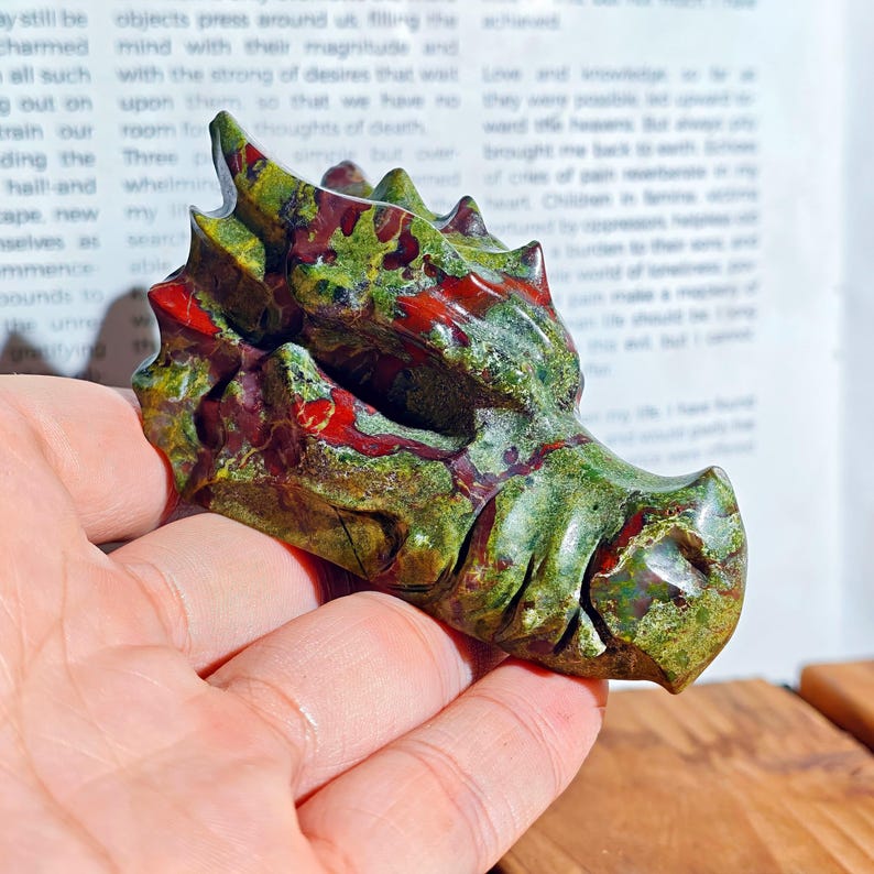 May include: A carved stone dragon head sculpture in shades of green and red. The dragon's detailed features include scales, horns, and a defined snout. The sculpture is held in a hand, with a blurred background.