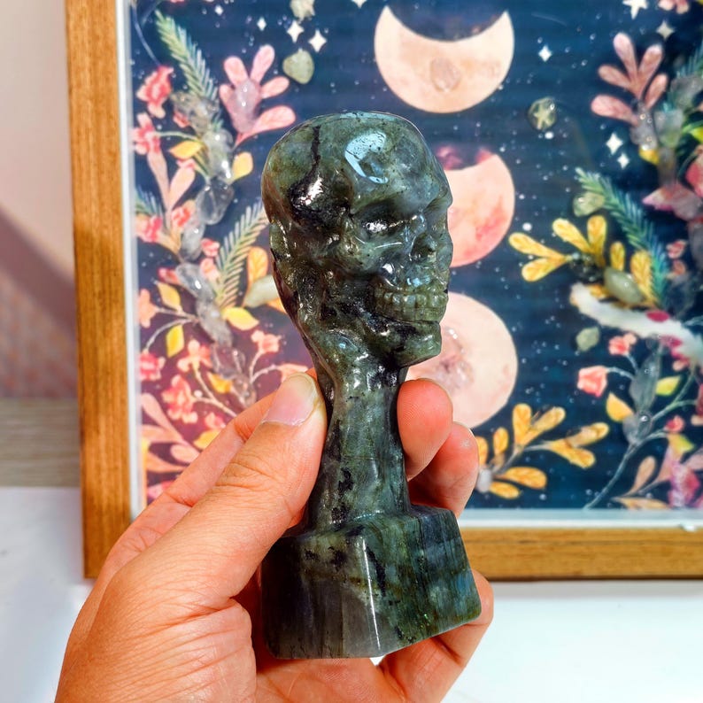Puede incluir: Una escultura de calavera tallada en labradorita, de color verde oscuro y azul. La calavera se sostiene en una mano, con un fondo decorativo con elementos florales y celestiales.