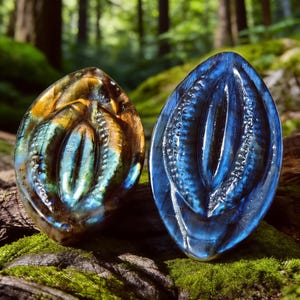 May include: Two carved labradorite gemstone pendants. One is a mix of gold, blue, and green, the other is a deep blue. Both have an oval shape with intricate carved details, set against a mossy background.