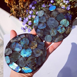 May include: Two round coasters with a mosaic design. The coasters are made of polished, oval-shaped stones in shades of blue, teal, and gray, set against a dark background. The stones have a shimmering, iridescent quality. The coasters are approximately 4 inches in diameter.