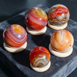 Puede incluir: Cinco esferas de ágata pulida en tonos rojos, naranjas y blancos, cada una sobre un pequeño soporte redondo de madera. Las esferas muestran patrones de bandas únicos y variaciones de color, creando una exhibición visualmente atractiva. El fondo es una superficie gris oscuro.