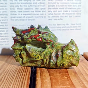May include: A carved dragon head sculpture in shades of green, red, and yellow, resting on a wooden surface. The dragon's detailed features include textured scales and sharp teeth. The sculpture is a decorative item.