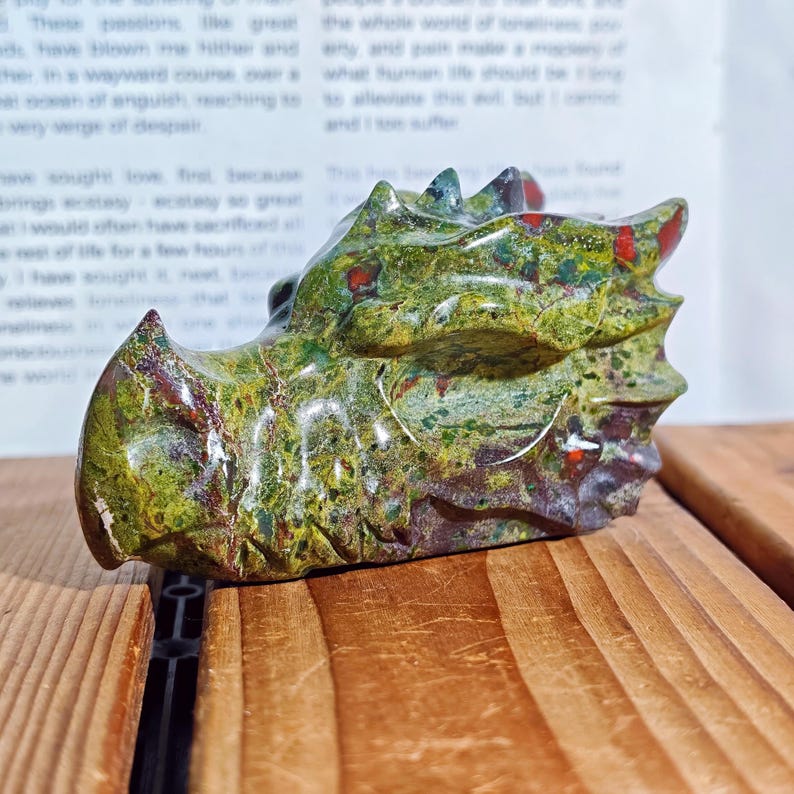 May include: A carved stone dragon head sculpture. The dragon is primarily green with red and brown accents. The sculpture is resting on a wooden surface.