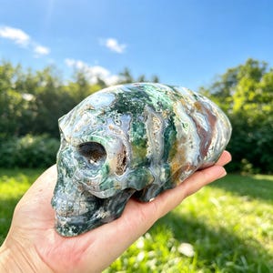 May include: A carved stone skull featuring a polished surface with intricate patterns of deep forest green, translucent white, and earthy brown inclusions throughout the mineral structure, showcasing natural crystalline formations and textures.