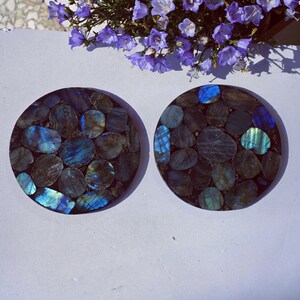 May include: Two round coasters with a mosaic design of blue, green, and gray stones. The stones are set in a dark brown base. The coasters are displayed on a white surface, with purple flowers in the background.