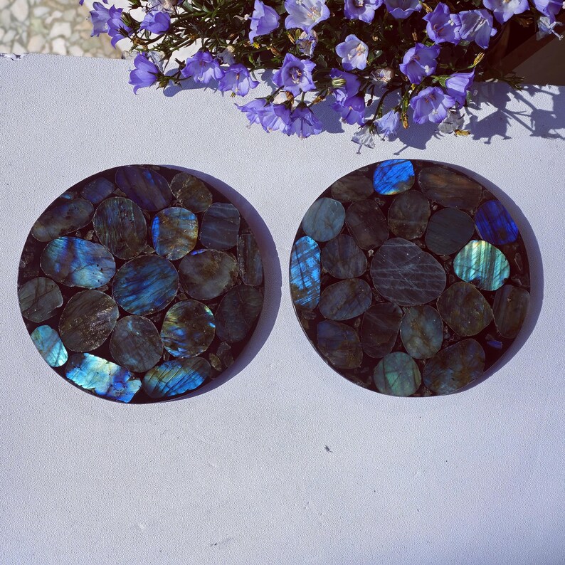 May include: Two round coasters with a mosaic design of blue, green, and gray stones. The stones are set in a dark brown base. The coasters are displayed on a white surface, with purple flowers in the background.