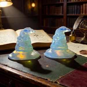 May include: Two opalescent glass wizard hats with a light source inside, casting a warm glow. The hats are on a dark green and red leather surface, with a vintage book and quill in the background. The hats are a light blue color.