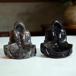 May include: Two stone sculptures of seated figures with hooded robes. One is a mottled gray and brown, the other is solid black. The figures are in a meditative pose, with their hands resting in their laps.