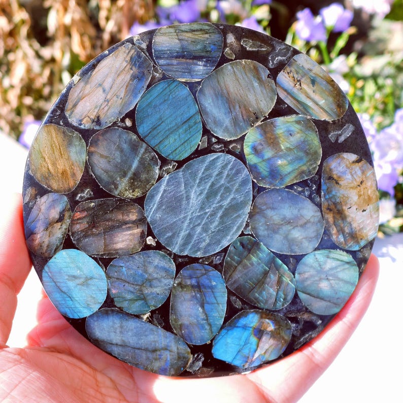 May include: A round, decorative coaster made of polished labradorite stones. The stones are various shades of blue, gray, and brown, set in a dark, possibly black, matrix. The coaster is held in a hand, with a blurred floral background.