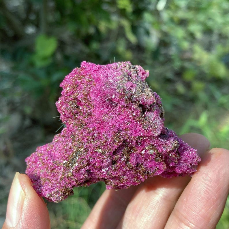 May include: A raw mineral specimen featuring a vibrant, saturated magenta-pink crystalline structure with embedded metallic bronze and gold-toned flecks throughout the rough, uneven surface, showcasing a natural, jagged, and porous geological formation.