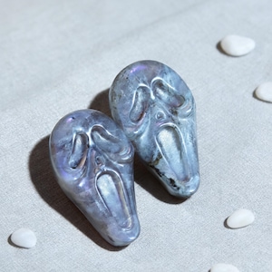 May include: Two carved, blue-grey gemstone pendants, each shaped like a screaming face. The pendants have iridescent qualities, with hints of purple and blue. Small white stones are scattered around the pendants on a light grey fabric.