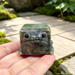 May include: A small, cube-shaped figurine carved from mottled dark green stone, featuring a flat top, a wide mouth, and two prominent, smooth, dark circular eyes. The object has a smooth, polished surface with natural veining throughout.