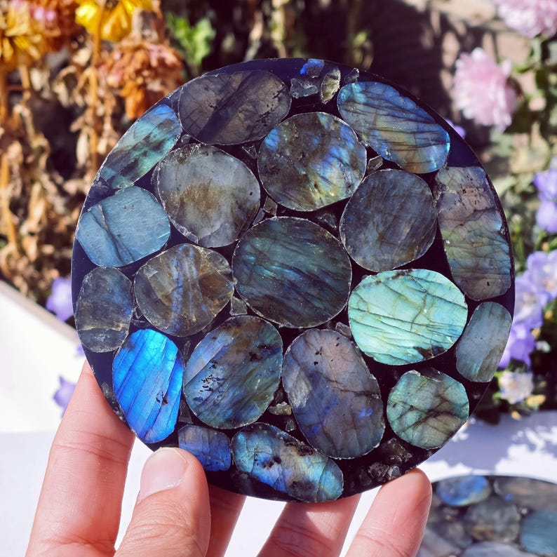 May include: A round coaster made of polished labradorite stones in shades of blue, teal, and gray. The stones are set in a dark, possibly black, resin base. The coaster is held by a hand, with flowers in the background.