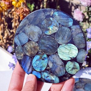 May include: A round coaster made of polished labradorite stones in shades of blue, teal, and gray. The stones are set in a dark, possibly resin, base. The coaster is held by a hand, with a blurred floral background.
