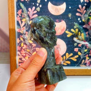 Puede incluir: Una escultura de cristal de labradorita tallada con forma de cabeza humana. La escultura es principalmente verde y negra con destellos iridiscentes. La cabeza se sostiene frente a una imagen enmarcada con un tema celestial, que incluye lunas y elementos florales.