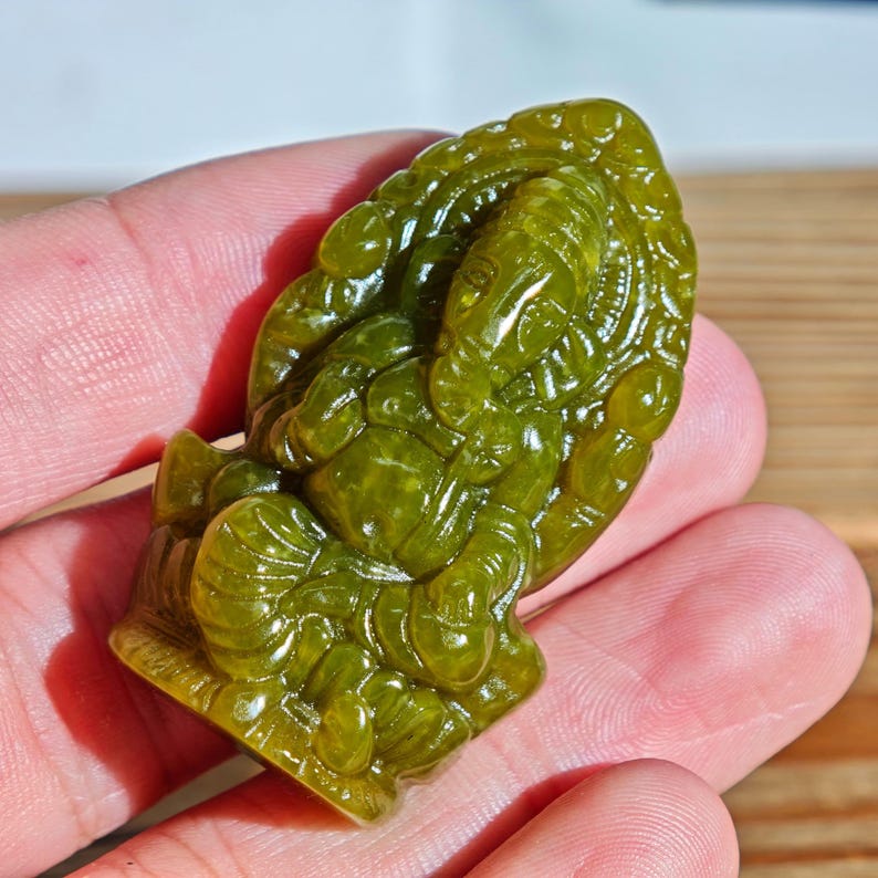 Puede incluir: Una detallada talla de jade verde del dios hind&uacute; Ganesha. La escultura se sostiene en una mano, mostrando los intrincados detalles del dios con cabeza de elefante. El jade tiene una calidad transl&uacute;cida, con variaciones en el color verde.
