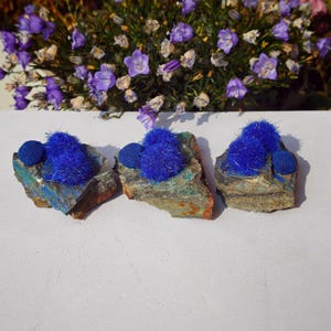 Puede incluir: Tres trozos de roca rugosa con vibrantes formaciones minerales azules. Cada roca tiene un grupo de cristales azules difusos y esf&eacute;ricos y una esfera azul m&aacute;s peque&ntilde;a y lisa. Las rocas est&aacute;n sobre una superficie blanca con flores moradas al fondo.