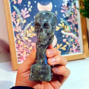 Puede incluir: Una figura de calavera tallada en piedra labradorita iridiscente. La calavera tiene detalles y est&aacute; montada sobre una base rectangular. La piedra tiene tonos grises, azules y verdes. El fondo incluye una impresi&oacute;n floral enmarcada.
