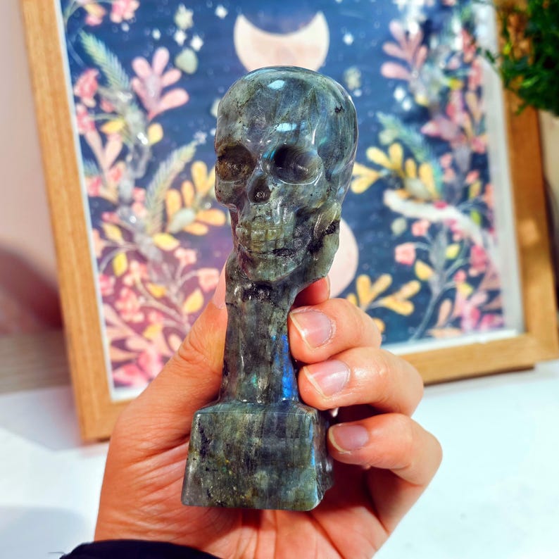 Puede incluir: Una figura de calavera tallada en piedra labradorita iridiscente. La calavera tiene detalles y est&aacute; montada sobre una base rectangular. La piedra tiene tonos grises, azules y verdes. El fondo incluye una impresi&oacute;n floral enmarcada.