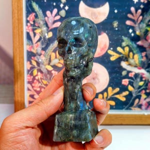 Puede incluir: Una figura de calavera tallada en piedra verde y azul iridiscente. La calavera tiene rasgos faciales detallados y se asienta sobre una base rectangular. El fondo presenta una impresi&oacute;n floral enmarcada con un tema celestial.