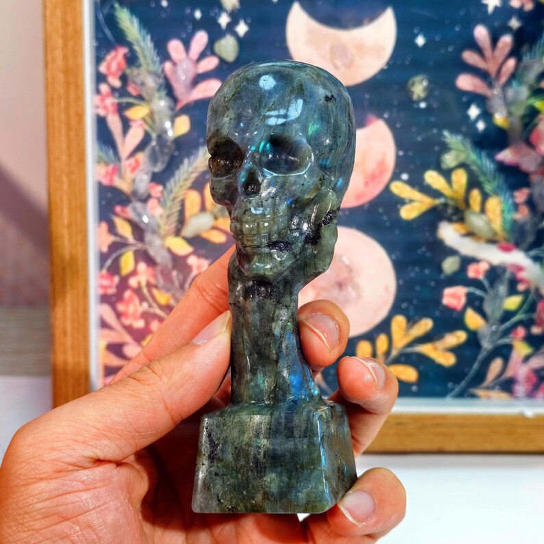 Puede incluir: Una figura de calavera tallada en piedra verde y azul iridiscente. La calavera tiene rasgos faciales detallados y se asienta sobre una base rectangular. El fondo presenta una impresi&oacute;n floral enmarcada con un tema celestial.