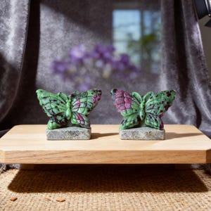 May include: Two carved butterfly sculptures. The butterflies are green with red and black accents, mounted on grey stone bases. They are displayed on a light brown wooden surface, against a grey backdrop.