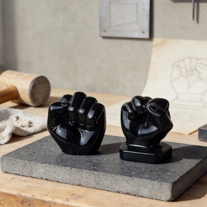 Könnte beinhalten: Zwei schwarze Faustskulpturen, eine davon auf quadratischem Sockel, stehen auf einer grauen rechteckigen Oberfläche. Die Skulpturen sind hochglanzpoliert und reflektieren das Licht. Der Hintergrund zeigt eine Handzeichnung und einen Holzhammer.