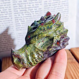 May include: A carved stone dragon head sculpture. The dragon is primarily green with red and purple accents. The sculpture has detailed scales and a textured surface, showcasing the craftsmanship of the piece. The dragon's head is held in a hand.