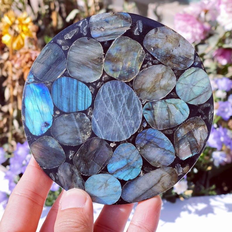 May include: Round coaster made of polished labradorite stones in shades of blue, gray, and teal, set in a dark matrix. The stones vary in size and shape, creating a mosaic-like pattern. The coaster is approximately 4 inches in diameter.