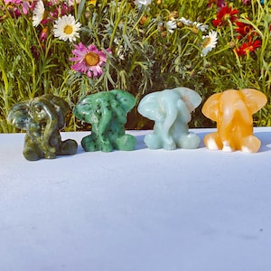 May include: Four small, carved elephant figurines in shades of green, blue, and orange. The elephants are sitting with their trunks raised. The background features a variety of colourful flowers.