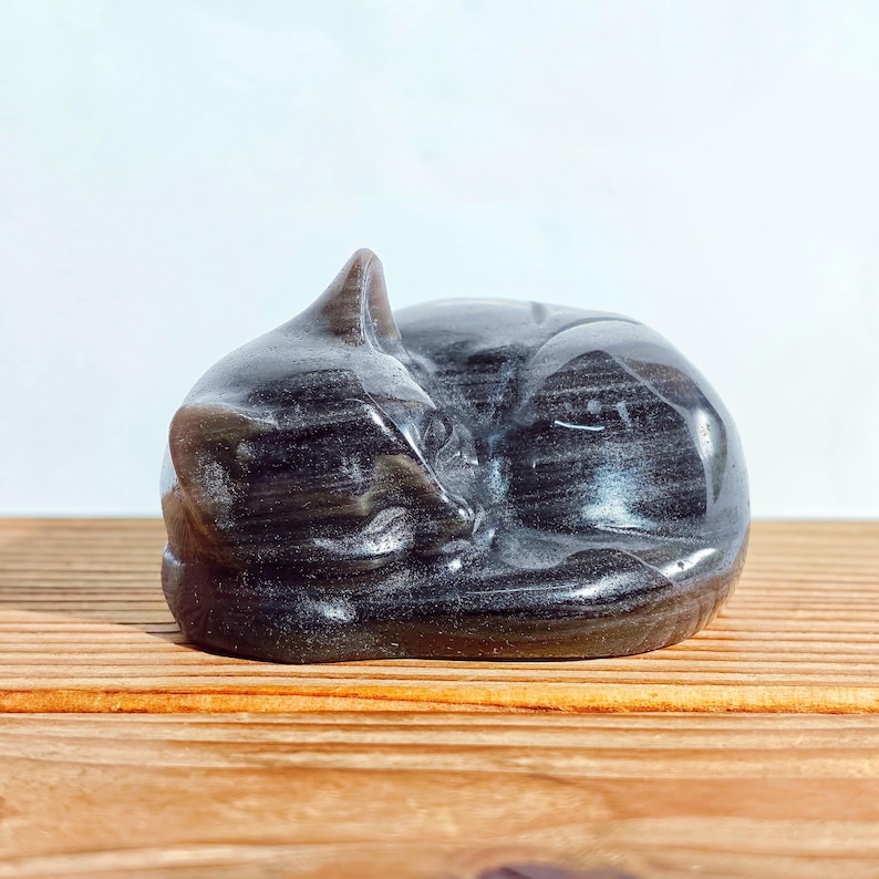 May include: A carved, sleeping cat figurine. The sculpture is a dark, swirling color, possibly obsidian or a similar stone. The cat is curled up, with its tail wrapped around its body. The figurine is resting on a wooden surface.