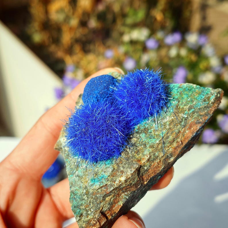 Puede incluir: Primer plano de un esp&eacute;cimen mineral con formaciones de cristales azul brillante sobre una roca multicolor. La roca muestra tonos verdes, marrones y azules, creando un contraste con los cristales azules brillantes. El esp&eacute;cimen se sostiene en una mano.