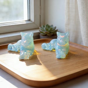 May include: Two iridescent cat figurines in a light blue hue are displayed on a wooden tray. The cats are in a relaxed pose, with their bodies elongated. The figurines have a glossy finish, reflecting light with a rainbow effect. A succulent plant is visible in the background.