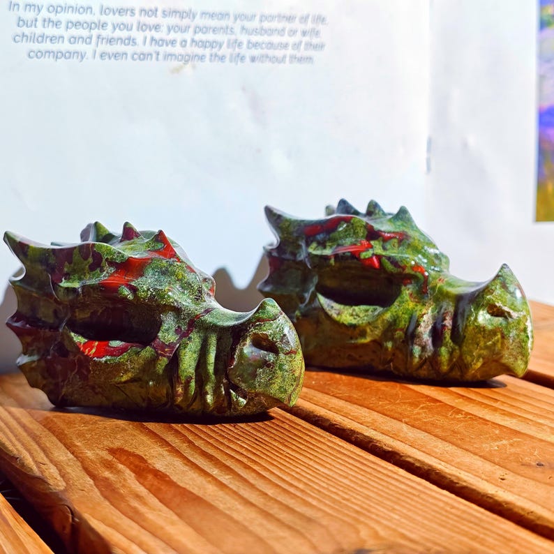 May include: Two carved dragon heads in green and red hues rest on a wooden surface. The dragon heads have detailed features, including scales and horns. The background includes text on a white page.