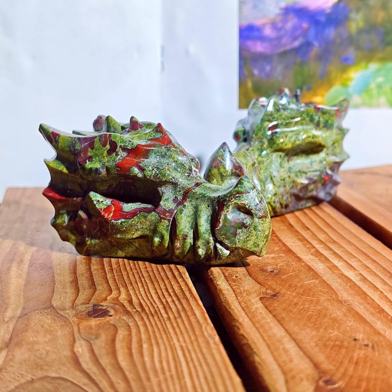 May include: Two carved stone dragon heads. The dragons are green with red accents. The heads are resting on a wooden surface. The dragon heads are detailed with scales and spikes.
