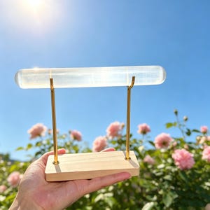 May include: A clear, cylindrical selenite crystal bar mounted horizontally on two thin, gold-toned metal rods. The rods are attached to a rectangular, light-coloured wooden base, creating a display stand for the translucent crystal piece.