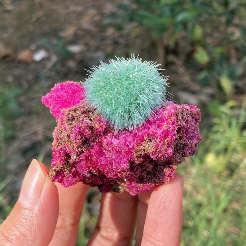 May include: A mineral specimen featuring a vibrant magenta, crystalline base with a textured, rocky surface. Attached to the top is a spherical, light green cluster of needle-like crystals radiating outward from a central point.