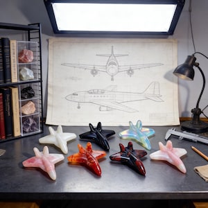 May include: A selection of decorative airplane ornaments in various colours, including white, black, red, and iridescent. The objects are displayed on a table with a blueprint of an aircraft in the background.