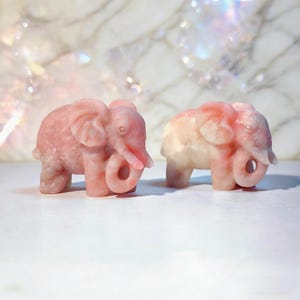 May include: Two carved elephant figurines. One is a solid pink color, and the other is a gradient of pink and white. The elephants have detailed trunks, ears, and tusks. The background is a white surface with a marble pattern.