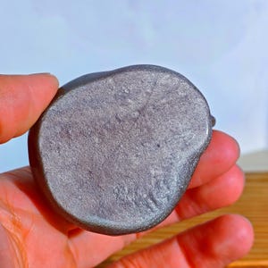 May include: A small, rounded, gray stone with a textured surface. The stone is held in a hand, showcasing its smooth, polished appearance. The stone's color is a muted gray with subtle variations.