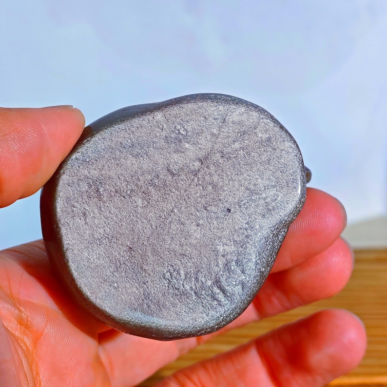 May include: A small, rounded, gray stone with a textured surface. The stone is held in a hand, showcasing its smooth, polished appearance. The stone's color is a muted gray with subtle variations.