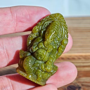 Puede incluir: Una figura tallada en piedra preciosa verde del dios hind&uacute; Ganesha. La escultura representa a Ganesha sentado, con detalles intrincados en la figura con cabeza de elefante y un fondo ornamentado. La piedra tiene un aspecto moteado.
