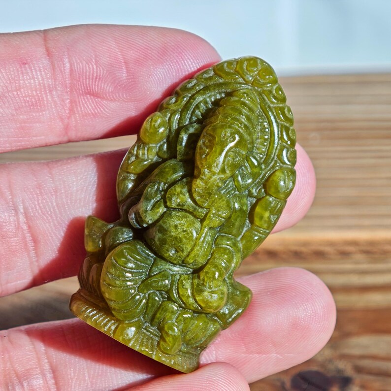 Puede incluir: Una figura tallada en piedra preciosa verde del dios hind&uacute; Ganesha. La escultura representa a Ganesha sentado, con detalles intrincados en la figura con cabeza de elefante y un fondo ornamentado. La piedra tiene un aspecto moteado.